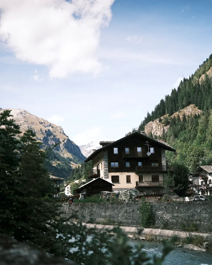 Casa di montagna lungo un torrente, con i boschi e le cime sullo sfondo